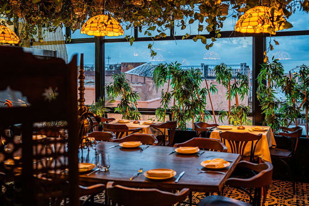 Chic restaurant interior featuring tables set for dining amid lush greenery, with cityscape visible through large windows.